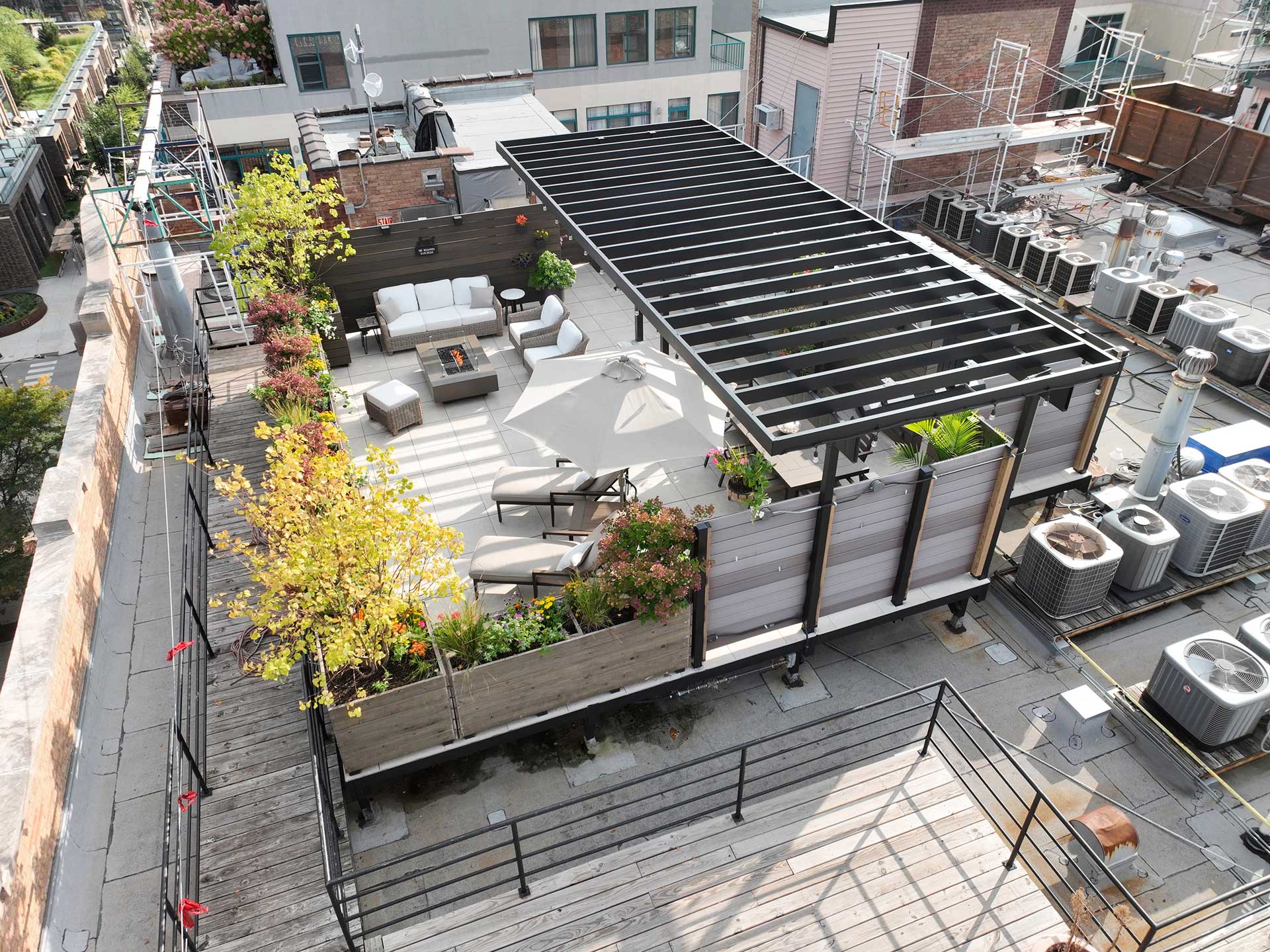 Aerial view of a spacious rooftop deck with a steel pergola, outdoor furniture, and lush planters